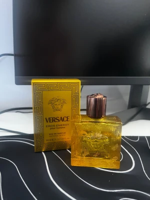 Versace Eros Energy Pour Homme parfym - Versace Eros Energy Pour Homme Eau de Parfum i en snygg, fyrkantig guldfärgad glasflaska med bronsfärgat lock. Flaskan har Versaces ikoniska Medusa-huvud och grekiskt mönster. Volymen är 50 ml och doften är för herrar.