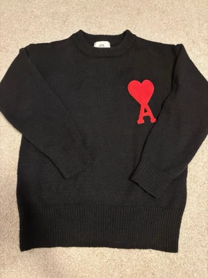 Svart stickad tröja Ami Paris - Säljer en svart stickad tröja från Ami Paris med deras ikoniska röda hjärta och bokstaven A broderat på bröstet. Tröjan har rund halsringning, långa ärmar och ribbade muddar. Perfekt för dig som gillar stilrena plagg med en twist.