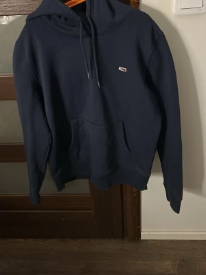 Mörkblå hoodie från Tommy Hilfiger - Snygg mörkblå hoodie från Tommy Hilfiger med klassisk huva och dragsnören. Har en liten broderad logga på bröstet och en stor magficka framtill. Tillverkad i mjukt bomullsmaterial som är skönt att ha på sig.