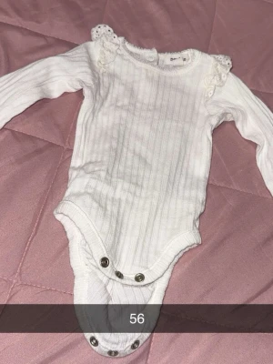 Vit ribbad body med spetsdetaljer - Söt vit långärmad body från Newbie med ribbad struktur och spetsdetaljer på axlarna. Tryckknappar i grenen för enkel påklädning. Perfekt för dig som gillar klassisk och gullig stil till de minsta.