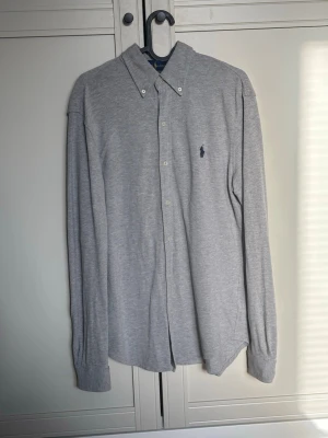 Grå skjorta från Ralph Lauren - Ralph Lauren skjorta | Klassisk grå skjorta från Ralph Lauren med button-down krage och broderad marinblå logga på bröstet. Skjortan har långa ärmar och knäppning framtill. Tillverkad i mjuk bomull för en bekväm känsla.