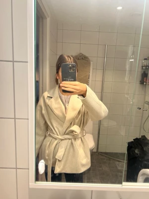 Beige kappa med knytskärp - Stilren beige kappa med bred krage och knytskärp i midjan. Kappan har en rak passform och är perfekt för lager-på-lager. Materialet ser ut att vara ullblandning och den har en klassisk look som funkar till många outfits.