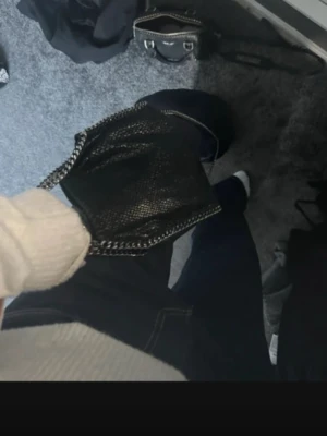 Svart Stella McCartney handväska - Säljer en svart handväska med kedjedetaljer runt kanterna och axelrem. Väskan har en mjuk, slouchy form och är gjord i veganskt läder med struktur som påminner om ormskinn. Perfekt för dig som gillar trendiga accessoarer.