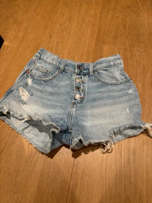 Blå denimshorts med knappar - Säljer ett par ljusblå denimshorts från Zara med relativ hög midja, slitna detaljer och fransig nederkant. Shortsen har fyra knappar framtill, klassiska fickor och en cool vintage-look. Perfekta för sommaren!