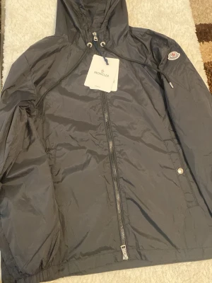 Svart vindjacka från Moncler - Snygg svart vindjacka från Moncler med huva, dragkedja framtill och logotyp på ärmen. Jackan har dragsko i huvan och knappdetaljer vid fickorna. Tillverkad i lätt och tåligt material som passar perfekt för blåsiga dagar.