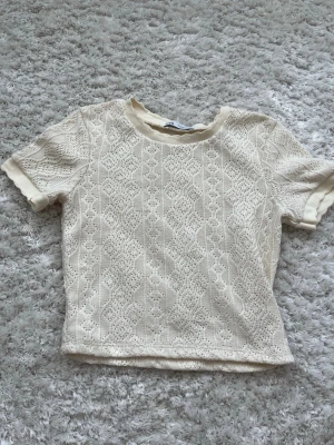 Vit stickad topp från Zara - Supersöt vit stickad topp från Zara med korta ärmar och rund hals. Blusen har ett detaljerat spetsmönster och ribbade kanter vid hals och ärmslut. Perfekt att styla med jeans eller kjol för en trendig look.
