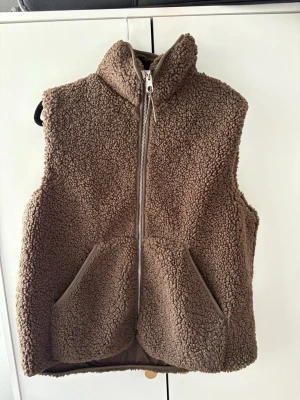Brun teddyväst med dragkedja - Mysig brun teddyväst i fluffigt material med hög krage och dragkedja framtill. Två stora fickor på framsidan ger en chill look. Perfekt att slänga över en hoodie eller tröja för extra värme nu till våren. Aldrig använd. 