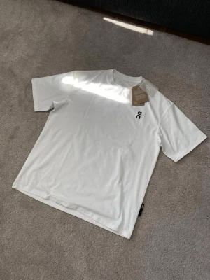 Vit t-shirt från On Clouds med logga - Säljer en stilren vit t-shirt från On med svart logga på bröstet och större logga på ryggen. Klassisk passform och rund halsringning. Perfekt till vardags för dig som gillar enkel och clean stil.