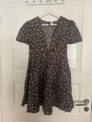 Svart blommig kortklänning - Söt svart kortklänning med små vita och beige blommor. Klänningen har v-ringning, korta ärmar och ett lätt utställt kjolparti. Perfekt för sommarens alla tillfällen och ger en avslappnad men trendig look.