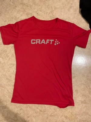 Röd träningströja från Craft 38 - Snygg röd träningströja från Craft med rund halsringning och korta ärmar. Tröjan har en enkel design med Craft-loggan i silver på bröstet. Tillverkad i ett lätt och snabbtorkande material som passar perfekt för träning och sportaktiviteter.