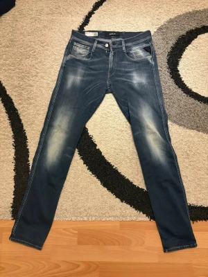 Blå slim fit jeans från Replay - Säljer ett par blå slim fit jeans från Replay med snygga slitningar och tvättade detaljer på låren och knäna. Klassisk femficksmodell med dragkedja och knapp. Jeansen har en modern passform och är tillverkade i bomull med lite stretch för extra komfort.