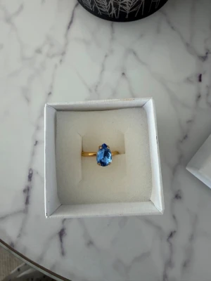 Caroline Svedbom - Snygg guldfärgad ring från Caroline Svedbom med en stor droppformad blå sten som mittpunkt. Ringen har en elegant och modern design som verkligen drar blickarna till sig. Perfekt accessoar för dig som vill sticka ut lite extra.