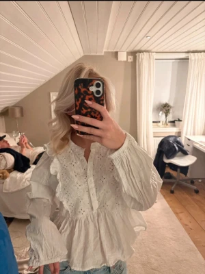 Vit volangblus med broderi - Säljer en söt vit blus med volanger och broderade detaljer. Blusen har lång ärm, knappar framtill och en luftig passform. Perfekt att styla med jeans eller kjol för en trendig look.