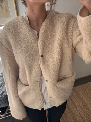 Beige teddyjacka med tryckknappar - Mysig beige teddyjacka med rund halsringning och tryckknappar framtill. Jackan har två stora fickor och en avslappnad passform. Aldrig andvänd har endast tagit av lapparna.