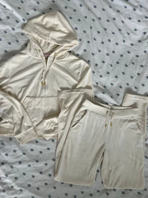 Krämvitt Juicy Couture loungewear-set - Säljer ett krämvitt / beige loungewear-set från Juicy Couture i mjuk velour. Setet består av en hoodie med dragkedja, fickor och guldfärgade detaljer samt matchande byxor med dragsko och fickor. Båda delarna har diskret JC-logga broderad framtill. Trojan är XL och byxorna är S. Kläderna är stretchiga och justerbara, midjebandet har dragsko som kan knytas. Hade passat S-L