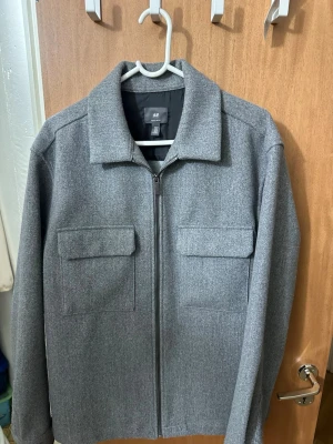 Grå overshirt från H&M med fickor - Stilren grå overshirt från H&M med rak passform och två stora bröstfickor med lock. Dragkedja framtill och klassisk krage. Tillverkad i ett mjukt, filtliknande material som ger en clean look. Perfekt att bära över en hoodie eller t-shirt.