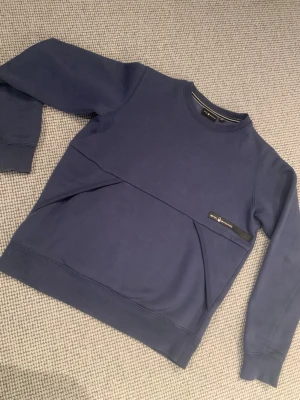 Blå sweatshirt från Sail Racing - Mörkblå sweatshirt från Sail Racing med rund halsringning och diskret logga på bröstet. Tröjan har en unik diagonal söm framtill och ribbade muddar vid ärmslut och nederkant. Perfekt för en avslappnad och stilren look.