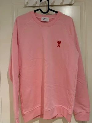 Rosa sweatshirt från Ami Paris - Storlek S, Säljer en ljusrosa sweatshirt från Ami Paris med deras ikoniska röda hjärtemblem broderat på bröstet. Tröjan har rund halsringning, långa ärmar och ribbade muddar. Perfekt för en chill och trendig look.