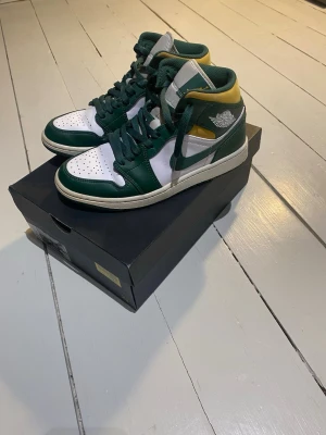Nike Air Jordan 1 High grön/vit/gul - Säljer ett par Nike Air Jordan 1 Mid Sonic sneakers i grönt, vitt och gult. Skorna har klassisk hög siluett, gröna snören och detaljer samt vit tåbox och sidor. Tillverkade i läder med ikoniska Jordan Wings-loggan på sidan och Swoosh i grönt.