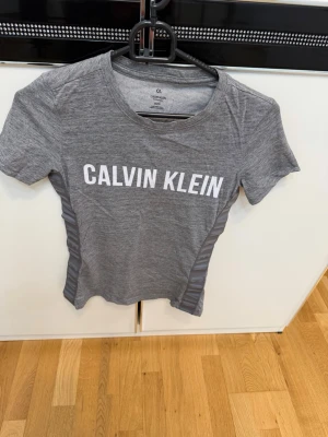 Grå Calvin Klein t-shirt - Snygg grå t-shirt från Calvin Klein med vit logga framtill. T-shirten har korta ärmar och är tillverkad i ett mjukt material som känns skönt mot huden. Passformen är slim och den har diskreta ränder på sidorna för en sportig look.