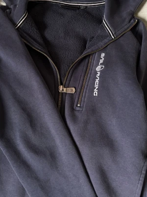 Marinblå hoodie från Sail Racing(pris diskuteras) - Oanvänd marinblå hoodie från Sail Racing med dragkedjaoch vit logga på bröstet. Huva med vita ränder på kanten och mjukt, fleecefodrat innerfoder. Perfekt för dig som gillar sportig och avslappnad stil.