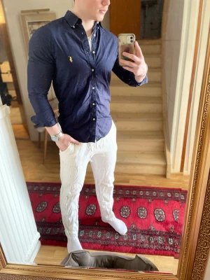 Mörkblå skjorta från Polo Ralph Lauren - Säljer en mörkblå skjorta från Polo Ralph Lauren i slim fit-modell. Skjortan har klassisk krage, långa ärmar och knappar framtill. Ikoniska Polo-loggan i gult på bröstet. Perfekt för dig som gillar stilrena och tidlösa plagg.