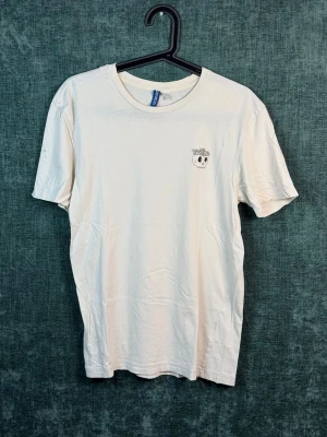 Vit t-shirt med döskalletryck - Vit t-shirt i bomull med ett litet svart tryck av en döskalle med blommor på huvudet på bröstet. Klassisk rund halsringning och korta ärmar. Perfekt för dig som gillar minimalistiska detaljer och vill ha en chill look.