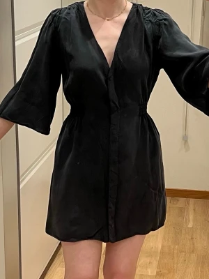 Svart kort omlottklänning zara  - Svart kort klänning från ZARA med v-ringning och omlottkänsla. Klänningen har korta ärmar, resår i midjan och är tillverkad i ett lätt tyg som ger en avslappnad look. Perfekt för dig som gillar enkel och stilren design.