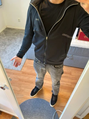 Svart hoodie från BOSS - Säljer en svart hoodie från BOSS med dragkedja framtill och två fickor. Diskret BOSS-logga på bröstet och ett mönstrat band över bröstet. Hoodien har huva och är tillverkad i mjukt material, perfekt för en avslappnad och stilren look. Den har en liten brun fläck vid boss märket