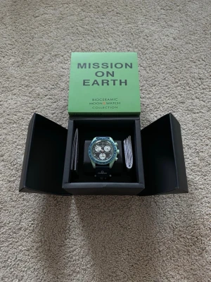 Omega x Swatch MoonSwatch Earth - Säljer en Omega x Swatch MoonSwatch Mission To Earth  från Bioceramic MoonSwatch Collection. Klockan har en mintgrön boett, blå bezel med tachymeter och svart urtavla med vita subdials. Snygg och unik design med texten 'Mission on Earth' på boxen. Levereras i originalförpackning.