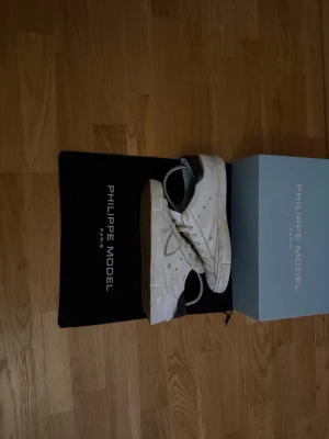 Vita Philippe Model sneakers - Stilrena vita sneakers från Philippe Model med svart hälparti och klassisk låg siluett. Skorna har vita snören, perforerade detaljer på sidan och är tillverkade i läder. Perfekta för dig som gillar en clean och tidlös look. Och boxen ingår ej 