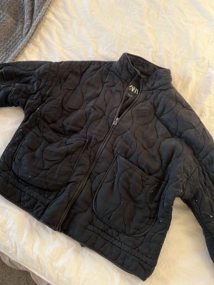 Svart quiltad pufferjacka från Zara - Säljer en svart quiltad pufferjacka från Zara med hög krage och dragkedja framtill. Jackan har två stora fickor och ett vågigt mönster i tyget. Perfekt för kalla dagar och riktigt snygg till streetwear-stilen.