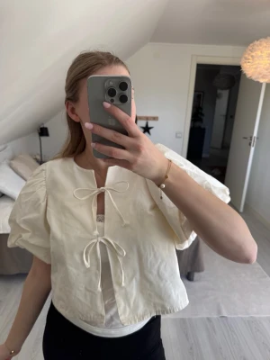 Vit blus med puffärmar och knyt - Trendig vit blus med stora puffärmar och två knytband framtill. Blusen har en rak passform och är tillverkad i ett lätt, vävt material som ger en luftig känsla. Perfekt för dig som vill ha en söt och stilren look med det lilla extra.