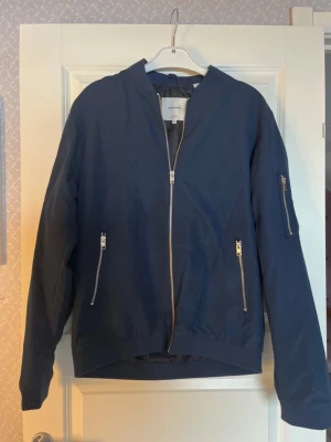 Mörkblå bomberjacka Jack & Jones - Snygg mörkblå bomberjacka från Jack & Jones med silvriga dragkedjor framtill och på ärmen. Jackan har klassisk bomberkrage, två fickor med dragkedja och är tillverkad i ett lätt material med fodrad insida. Perfekt för dig som vill ha en stilren och enkel look.