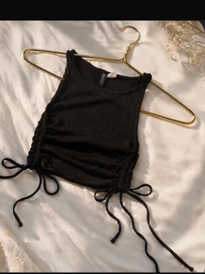 Svart croppad linnetopp från Divided - Snygg svart croppad linnetopp från Divided med snörning i sidorna. Toppen är ärmlös och har en tight passform som framhäver midjan. Perfekt att matcha med höga jeans eller kjol för en trendig look.