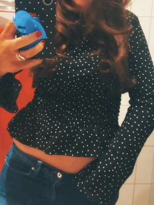 Svart prickig blus med smock - Trendig svart blus med vita små prickar och smockad midja som ger en snygg siluett. Blusen har långa ärmar med lätt utsvängd form och volang nedtill. Tillverkad i ett lätt och luftigt tyg, perfekt att styla med jeans eller kjol.