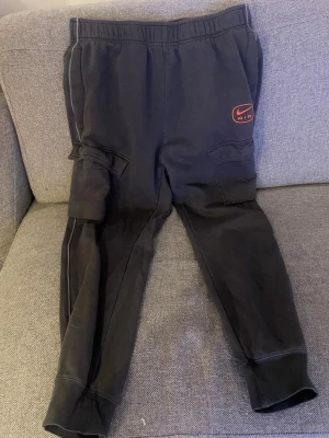 Mörk grå Nike Air mjukisbyxor cargo - Mörkgrå mjukisbyxor från Nike Air med cargo-fickor på sidorna och elastisk midja. Byxorna har en röd Nike Air-logga på vänster ben och är tillverkade i mjukt fleecematerial för extra komfort. Perfekta för en avslappnad och sportig stil.