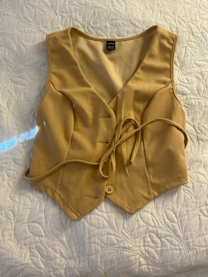 Beige omlottväst med knyt - Trendig beige väst/top  med omlottmodell och knytband i midjan. Topen har v-ringad hals och en knappdetalj framtill. 
