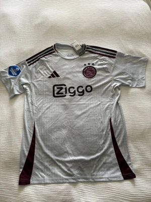 Ajax bortatröja 23/24 Bergwijn #7 - Ajax bortatröja säsongen 23/24 från Adidas med Ziggo som sponsor. Tröjan är vit med vinröda detaljer, klubbmärke och Adidas-logga på bröstet samt Bergwijn och nummer 7 på ryggen. Tillverkad i lätt och ventilerande material, perfekt för fotbollsträning eller match.