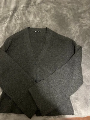 Mörkgrå kofta med v-ringning - Mysig mörkgrå kofta med v-ringning och två knappar framtill. Koftan har lång ärm och är tillverkad i ett mjukt, stickat material som passar perfekt för lager på lager. Enkel och stilren design som funkar till det mesta.