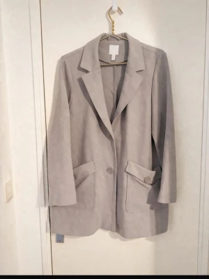 Ljusgrå oversized kavaj - Stilren ljusgrå kavaj med oversized passform och två stora fickor framtill. Kavajen har klassisk krage, två knappar och är tillverkad i ett mjukt material som påminner om mocka. Perfekt för en trendig och avslappnad look.