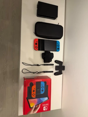 Nintendo Switch OLED med tillbehör - Nintendo Switch OLED med neonröda och neonblå Joy-Con, svart dockningsstation, svart bärväska, handkontrollhållare, handledsremmar och originalkartong. Tre spel ingår: Super Mario Odyssey, Nintendo Switch Sports och Pokémon Scarlet.