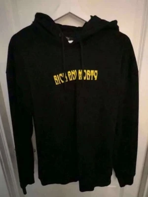 Rick and Morty svart hoodie - Svart hoodie med huva och snörning, prydd med gult Rick and Morty-tryck på bröstet och en stor färgglad grafik på ryggen med karaktärerna från serien. Perfekt för dig som älskar animerade serier och vill sticka ut med en unik och cool design.