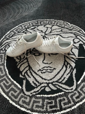 Vita Fred Perry sneakers i läder - Snygga vita sneakers från Fred Perry i klassisk låg modell. Skorna är tillverkade i slätt läder med vita snören och diskret logga på plösen. Perfekta för dig som gillar stilrena och tidlösa sneakers till vardagslooken.