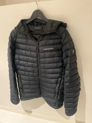 Svart dunjacka Peak Performance  - Snygg svart dunjacka från Peak Performance med huva och dragkedja framtill. Jackan har diskret logga på bröstet och vänster ärm, quiltad design och två sidofickor. Perfekt för kalla vinterdagar och riktigt lätt att bära.