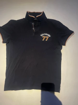 Svart Sail Racing t-shirt med half zip - Snygg mörkblå t-shirt från Sail Racing med korta ärmar och knappar vid halsen. Tryck och orange/vita detaljer på krage och ärmslut. Perfekt för en sportig och avslappnad stil.