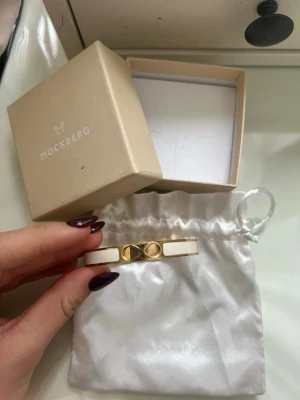Mockberg Armband  - Snyggt armband från Mockberg i vitt och guldigt metall. Armbandet har en elegant design med vita detaljer och guldfärgade inslag, samt en dekorativ mittdel. Perfekt accessoar för att lyfta din outfit. Helt oanvänt och inga decken på använing. Äkta såklart!!