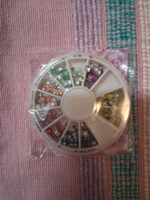 Mixade nageldekorationer i hjul - Säljer ett runt plastfodral fyllt med mixade nageldekorationer i olika färger och former, som pärlor, stenar och nitar. Perfekt för att skapa unika nageldesigner och pynta naglarna med färgglada detaljer.