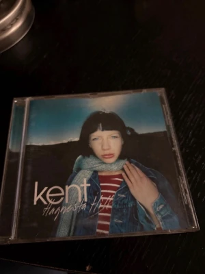 Kent Hagnesta Hill CD-album - Säljer Kent-albumet Hagnesta Hill på CD i original plastfodral. Omslaget har ett foto med blå himmel och person i jeansjacka och randig tröja. Skivan är vit med tryckt text och RCA-logga. Perfekt för dig som gillar svensk alternativ rock.
