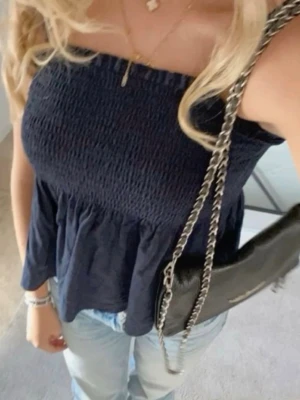 Mörkblå tubtopp med volang - Trendig mörkblå tubtopp med smockad överdel och volang nedtill. Toppen är axelbandslös och har en avslappnad passform. Perfekt till jeans eller kjol för en cool och enkel look. Materialet känns lätt och luftigt, troligen bomull eller viskos.